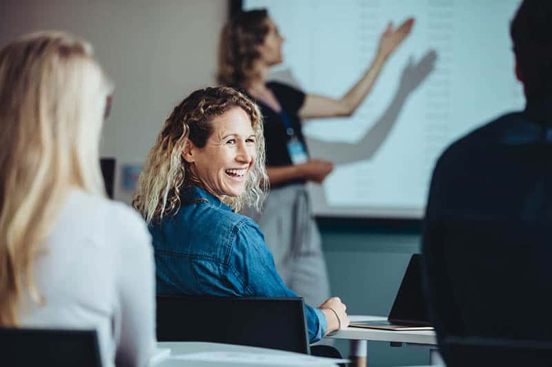 Smilende kvinne deltar i et kurs om GDPR og sikker lagring av personopplysninger, med en instruktør som forklarer regler og prosedyrer i bakgrunnen.