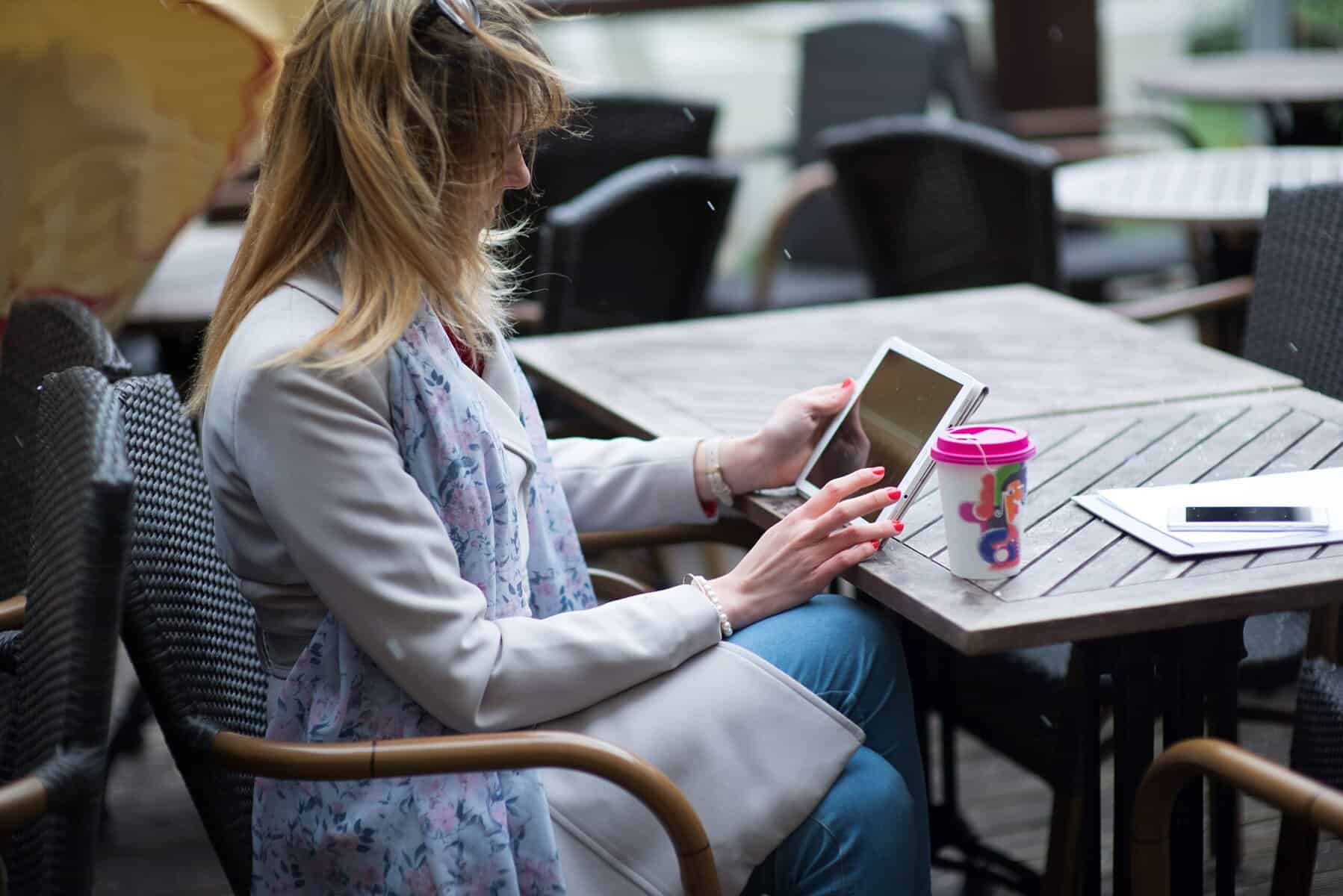 Kvinne sitter ved et kafebord med en takeaway kaffe og gjennomfører et kurs på nettbrettet sitt