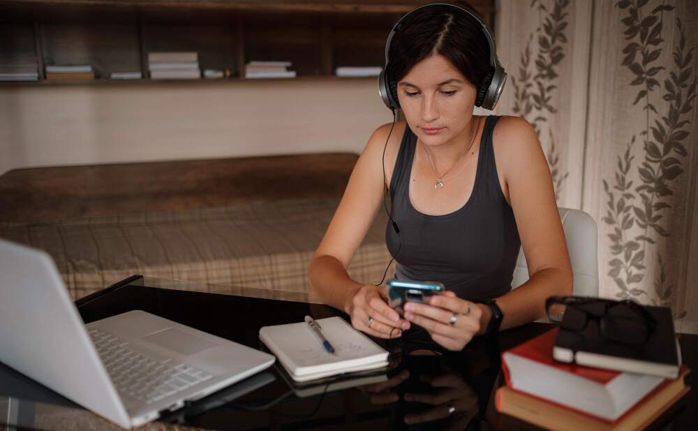 Kvinne sitter ved skrivebord og gjennomfører et kurs på telefonen med en laptop og notatbok liggende på bordet