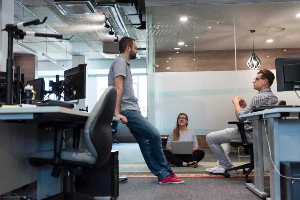 Kolleger diskuterer et emne på kontor, hvor den ene mannen står oppreist, den andre sitter i en stol og kvinnen sitter på gulvet med en laptop på fanget