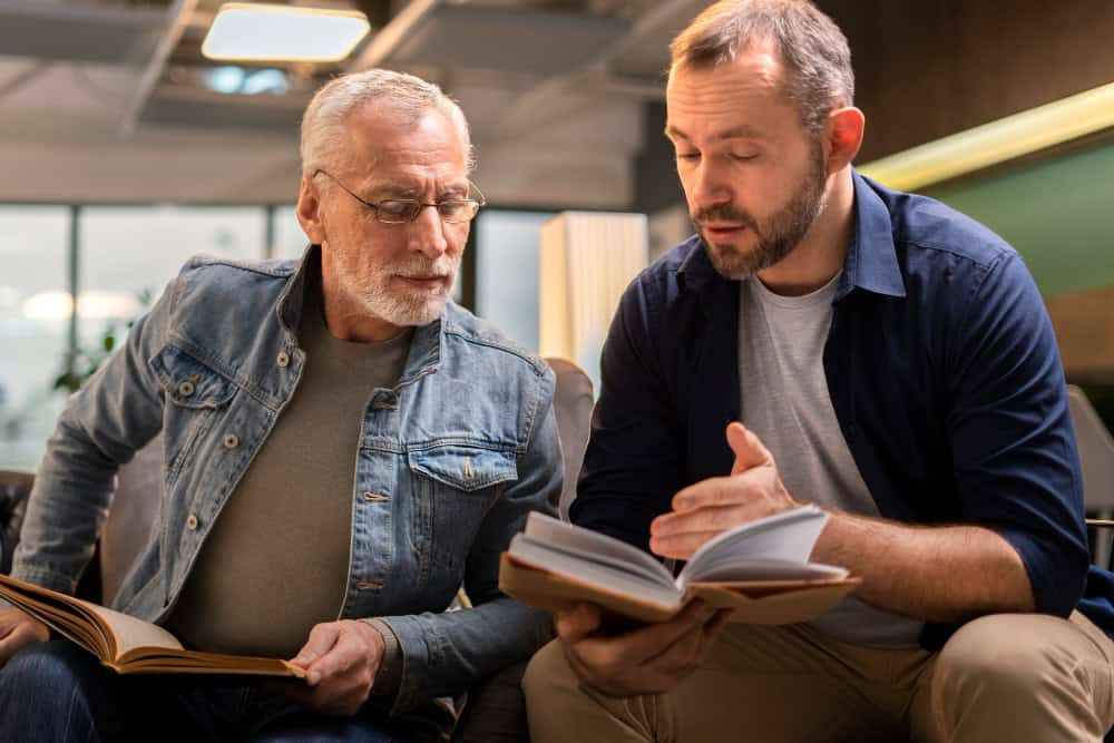 To menn sitter ved siden av hverandre og diskuterer en bok som den ene av dem leser