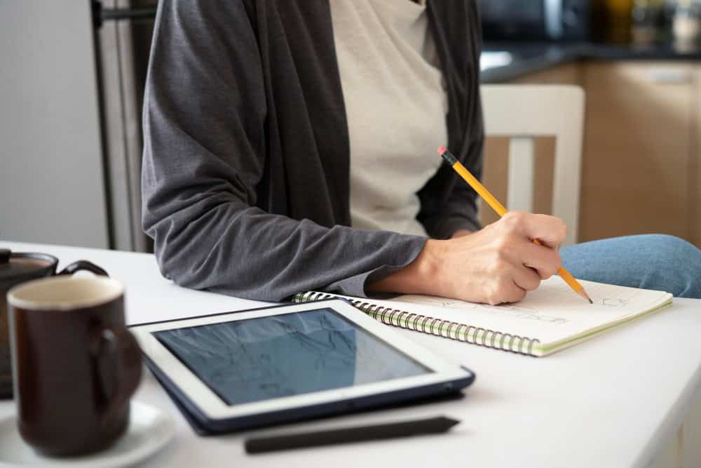 Kvinne sitter ved spisebordet sitt med en kopp kaffe og en iPad foran seg mens hun noterer i en skriveblokk